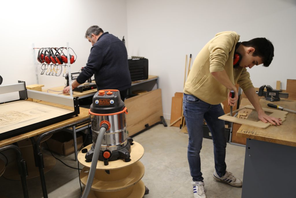 Deux personnes travaillent dans un atelier. L'une utilise une scie, l'autre une perceuse sur des planches de bois. Des écouteurs sont accrochés au mur et un aspirateur est au centre. L'espace est organisé avec des outils et des établis.