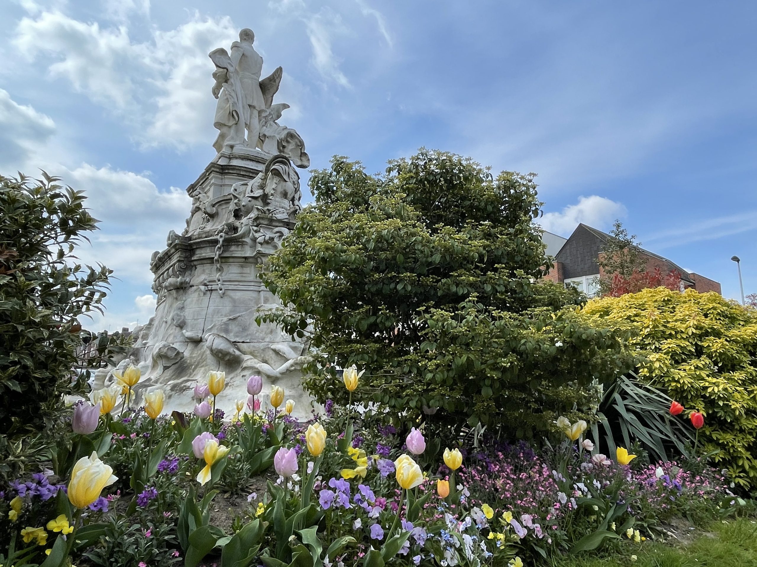 Un jardin luxuriant avec des tulipes colorées et diverses plantes entoure un grand monument en pierre finement sculpté contre un ciel bleu avec des nuages épars.