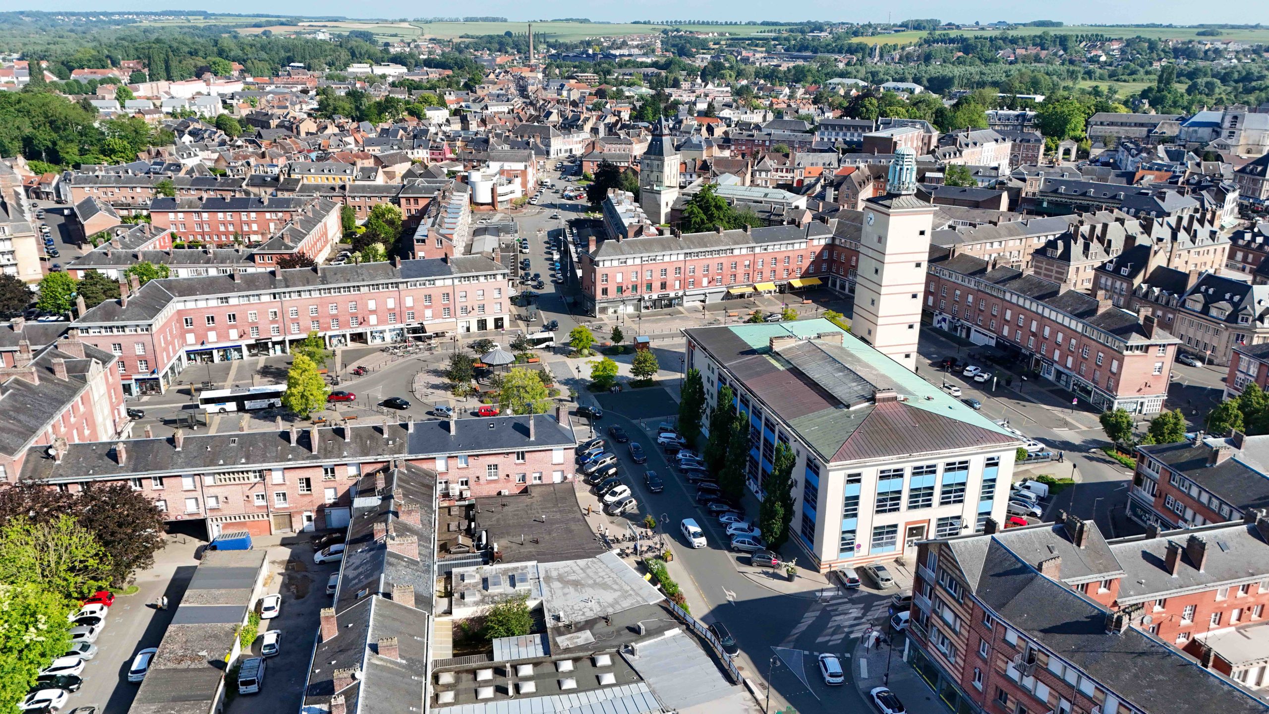 Vue aérienne d'une ville européenne avec son grand hôtel de ville rectangulaire surmonté d'une haute tour d'horloge. Tout autour, des rangées de bâtiments en briques rouges, des rues bordées de voitures garées et des paysages verdoyants au loin.