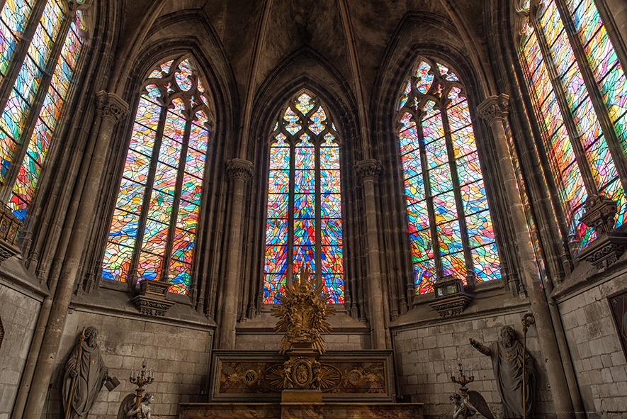 Intérieur d'une église comportant un grand autel avec des statues religieuses complexes, entouré de grands vitraux colorés représentant divers motifs abstraits, projetant une lumière vibrante sur les murs et le sol en pierre.