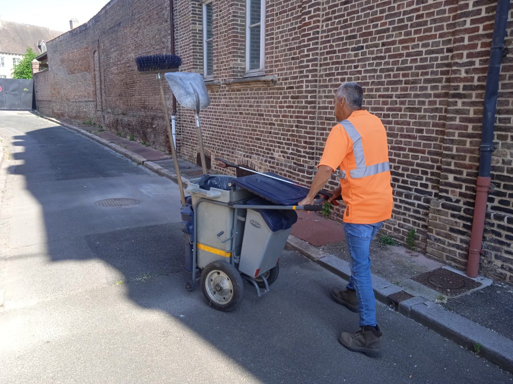 Une personne portant un gilet orange haute visibilité pousse un chariot de nettoyage dans une rue étroite bordée de murs en briques. Le chariot transporte un balai et une pelle à poussière. La rue est vide et le soleil projette des ombres sur le sol.