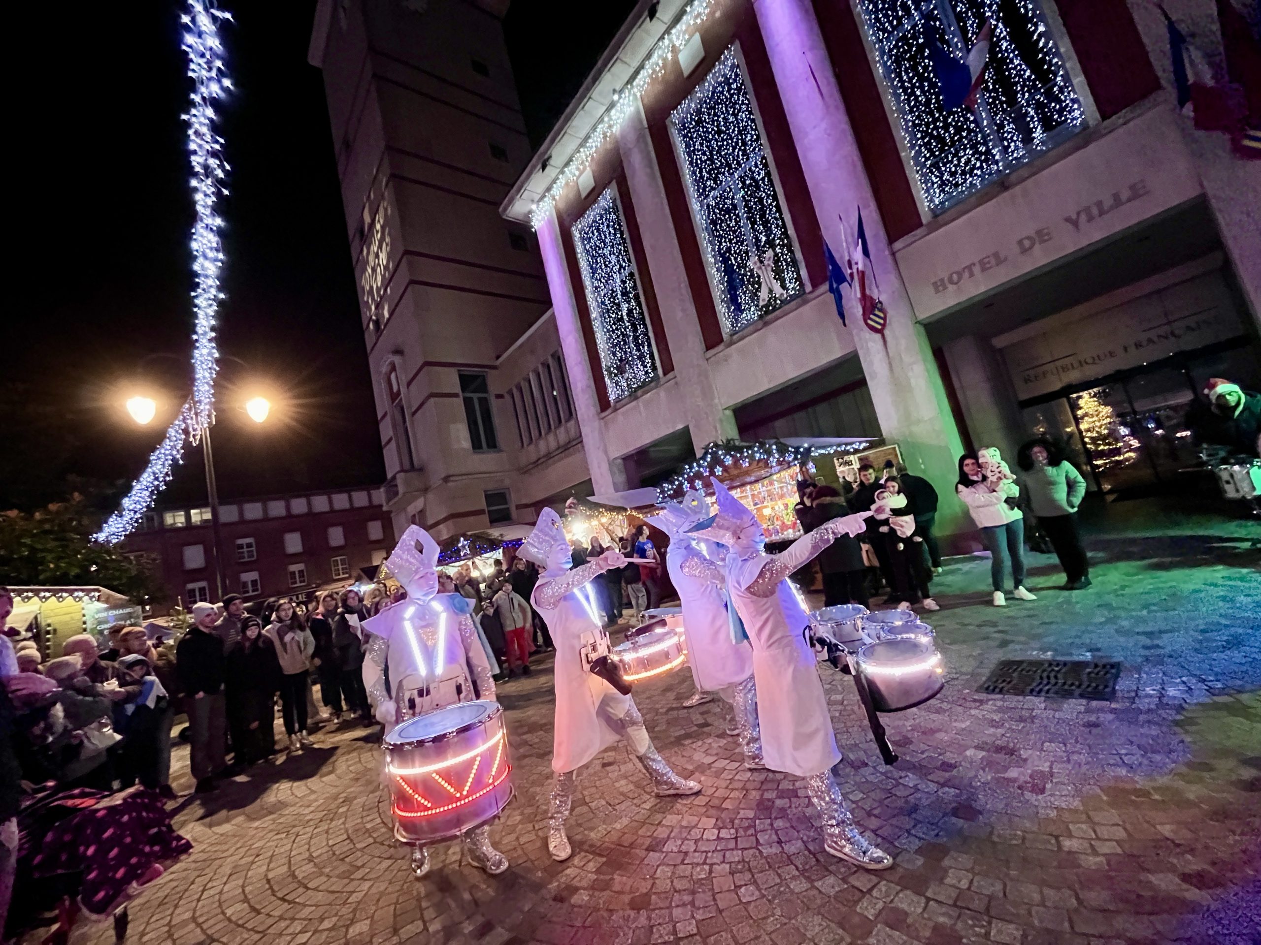 Un groupe d'artistes en costumes blancs avec des tambours illuminés joue devant un bâtiment illuminé. Les gens regardent le spectacle dans la rue pavée. Le bâtiment est décoré de lumières et un panneau indique « Hôtel de Ville ».