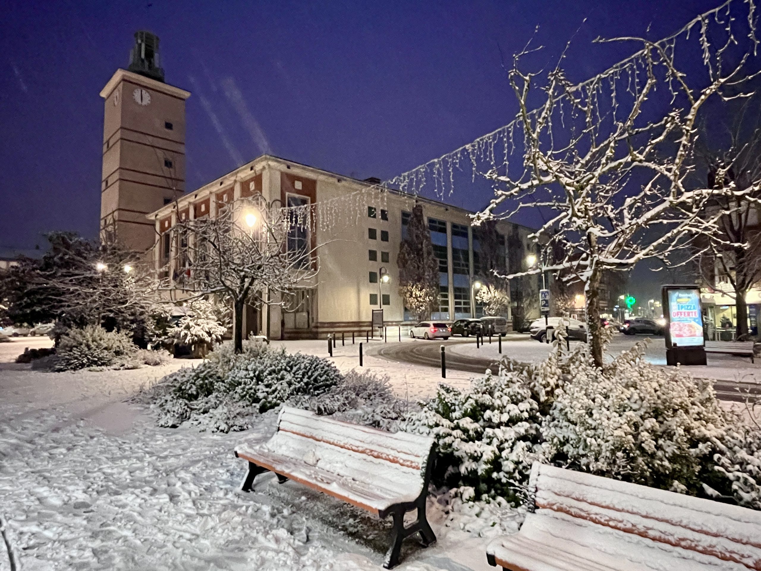 Une scène urbaine enneigée la nuit avec une haute tour d'horloge à gauche. Le sol et les bancs au premier plan sont recouverts de neige. Des arbres nus décorés de guirlandes lumineuses encadrent la scène et quelques voitures garées sont visibles à l'arrière-plan.