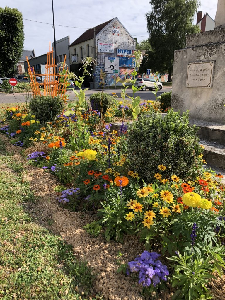 Un jardin animé avec diverses fleurs en fleurs jaunes, orange et violettes. Des buissons et quelques plantes plus hautes ajoutent de la profondeur. Un panneau en pierre est partiellement visible à droite. Une route et des bâtiments sont en arrière-plan sous un ciel clair.