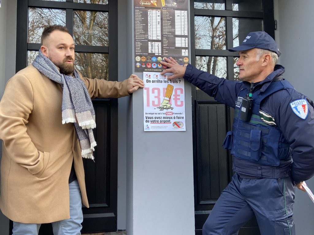 Un homme en manteau beige discute avec un policier en uniforme à l'extérieur d'un bâtiment. Ils discutent d'une affiche sur le prix des cigarettes, sur laquelle est écrit "13€" bien en évidence. Le policier pointe du doigt l'affiche, et en arrière-plan, on voit une porte avec un menu au-dessus.